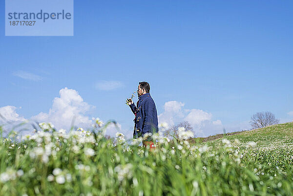 Seitenansicht eines Mannes  der auf einer Wiese steht und Blumen vor blauem Himmel duftet