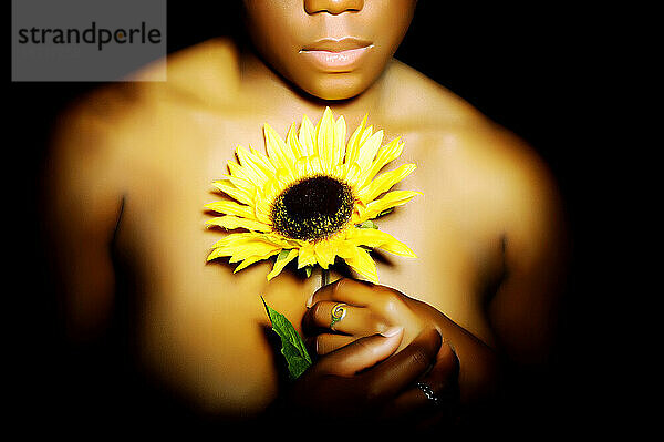 Eine junge afroamerikanische Frau bedeckt ihre nackten Brüste  indem sie eine Sonnenblume vor sich hält.