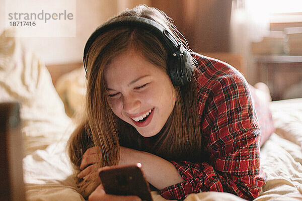 Glückliche junge Frau mit Kopfhörern und Smartphone im Bett liegend