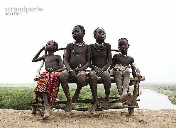 Vier kleine Jungen sitzen auf einer Bank mit Blick auf den Omo-Fluss im abgelegenen Omo-Tal in Äthiopien.