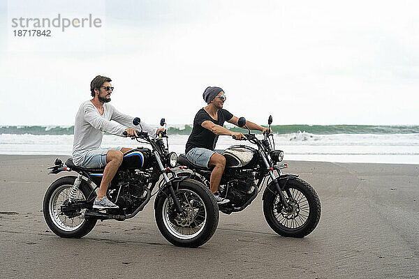 Zwei junge Männer fahren mit einem Retro-Motorrad am Strand.