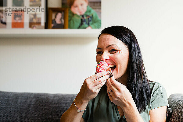 Eine Frau lacht  während sie zu Hause einen rosa Cupcake isst