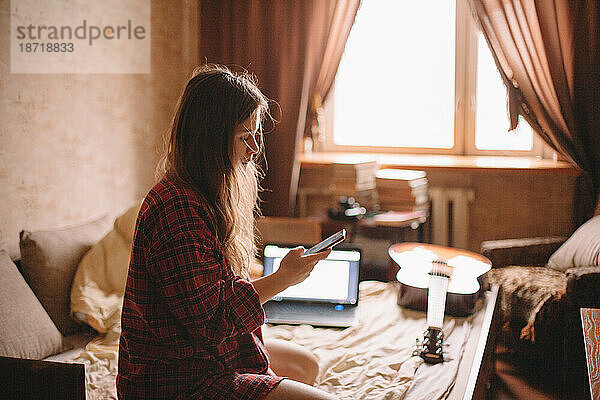 Junge Frau benutzt Smartphone  während sie zu Hause im Bett sitzt