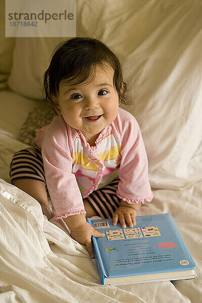 Ein glückliches kleines Mädchen sitzt mit einem Buch auf einem Bett  umgeben von weichen weißen Laken und lächelt in die Kamera.