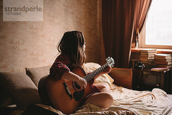 Junge Frau spielt Gitarre  während sie zu Hause im Bett sitzt