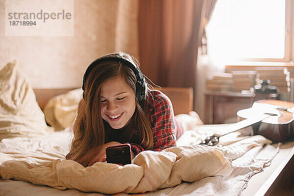 Fröhliches Teenager-Mädchen mit Kopfhörern und Smartphone im Bett liegend
