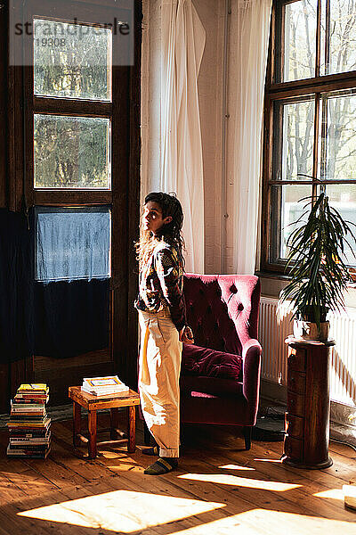 Frau steht zu Hause mit Pflanzen und Büchern im großen Fensterlicht
