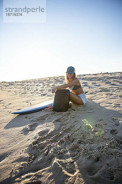 Frau sitzt bei Sonnenuntergang am Strand mit Surfbrett und Rucksack