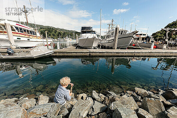 Vorschulkind sitzt auf Felsen in der Nähe von Booten in einem Hafen