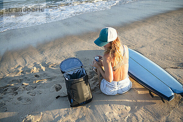 Frau entspannt sich bei Sonnenuntergang am Strand mit Surfbrett und Rucksack