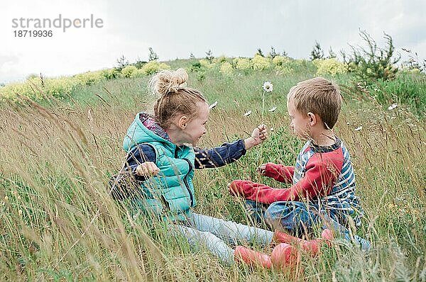 Ein Mädchen reicht einem Jungen eine Blume  während es mitten auf einem Feld sitzt