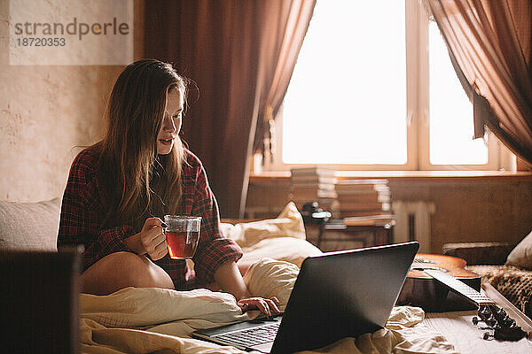 Junge Frau hält eine Tasse Tee in der Hand  während sie im Bett einen Laptop benutzt