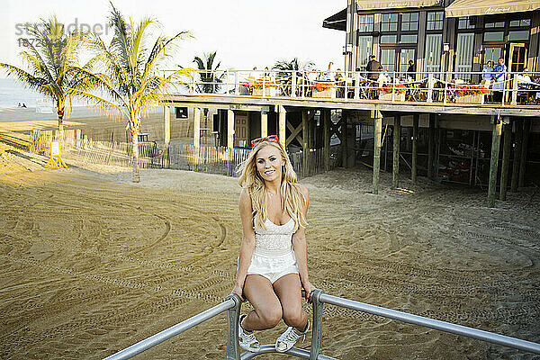 Blonde Frau sitzt auf Treppengeländer mit Blick auf den Strand