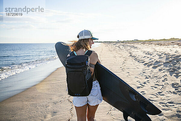 Frau spaziert bei Sonnenuntergang mit Surfbrett und Rucksack am Strand entlang