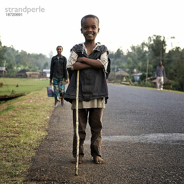 Ein Junge mit einem Spazierstock posiert am Rand einer gepflasterten Straße  während Dorfbewohner hinter ihm die Straße entlanggehen.