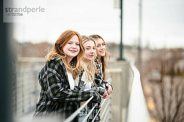 Drei glückliche Teenager-Mädchen lehnen sich draußen an einen Zaun.