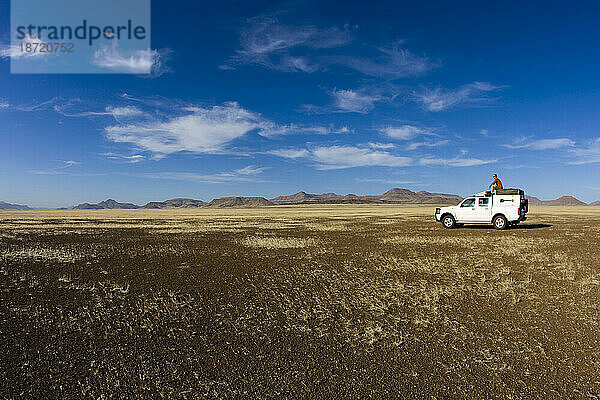 Mann sitzt auf dem Dach eines Expeditionslastwagens mitten in der afrikanischen Wüste. Weite Weite und blauer Himmel. Horizontales Bild.