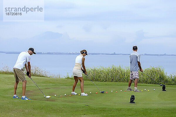 Drei Personen üben das Schlagen von Golfbällen mit Blick auf das Meer im Hintergrund