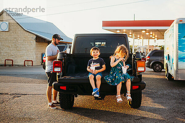Junge und Mädchen sitzen auf der Ladefläche eines Jeep-Lastwagens und essen mit ihrem Vater Schneekegel