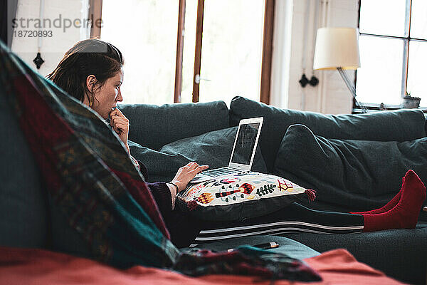 Frau sitzt auf einer gemütlichen Couch und arbeitet von zu Hause aus auf einem Laptop