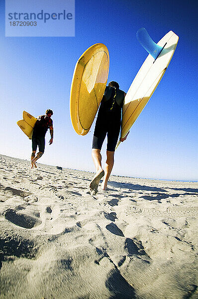 Die frühe Morgensonne scheint auf zwei junge Surfer  die Surfbretter den Strand entlang tragen.