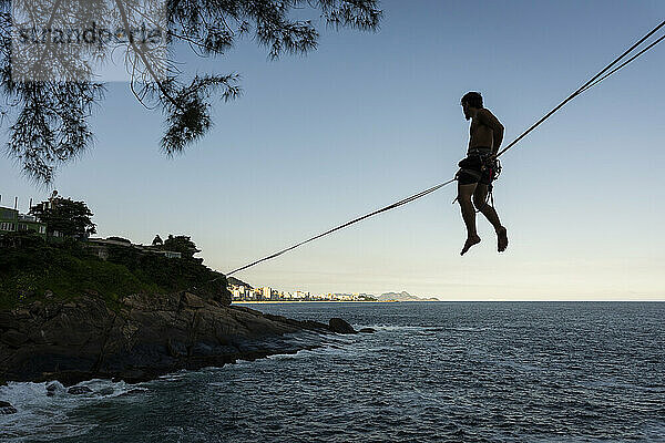 Silhouette eines Mannes auf einer Highline über dem Meer bei Sonnenuntergang