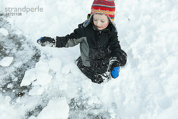 Kleiner Junge kniet nach Schneesturm spielerisch im Schnee und macht Schneebälle