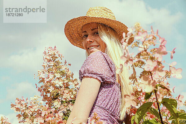 Lächelnde junge blonde Frau mit Strohhut in der Nähe von Hortensienblüten