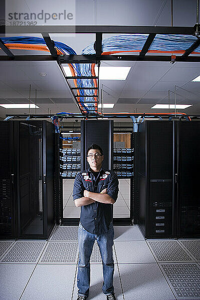 Ein junger erwachsener asiatischer Mann steht selbstbewusst vor einer Reihe neuer Netzwerkserver in einem Serverraum.