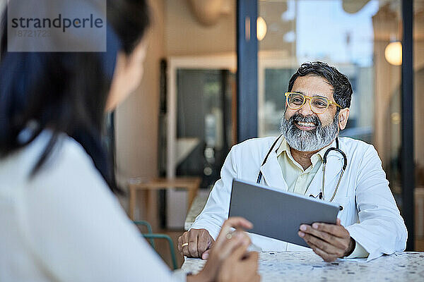Glücklicher Arzt hält Tablet-PC in der Hand  während er mit dem Patienten in der Klinik spricht