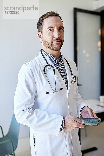 Porträt eines männlichen Arztes  der sein Smartphone in der Hand hält  während er in der Klinik steht