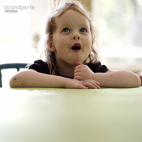 Ein kleines Mädchen mit blonden  lockigen Haaren macht ein überraschtes Gesicht  während es an einem Tisch sitzt.