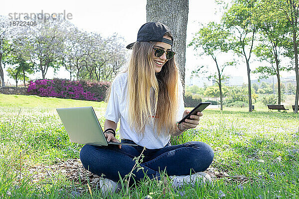 Vorderansicht einer glücklichen Studentin  die mit Laptop und Smartphone in einem grünen Park eines Universitätscampus arbeitet