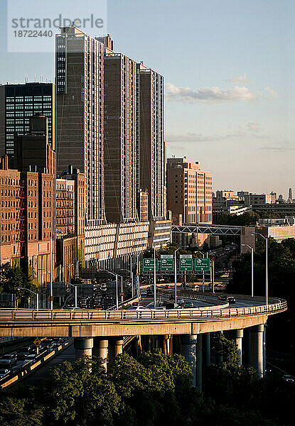 Eine erhöhte Autobahnauffahrt erstreckt sich vor einer Reihe hoher Gebäude  die von der untergehenden Sonne beleuchtet werden.