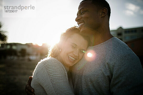 Multi racial couple embrace and laugh on beach at sunset