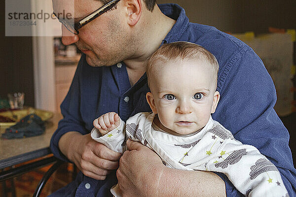 Ein Baby mit leuchtenden Augen sitzt auf dem Schoß seines Vaters an einem Esstisch
