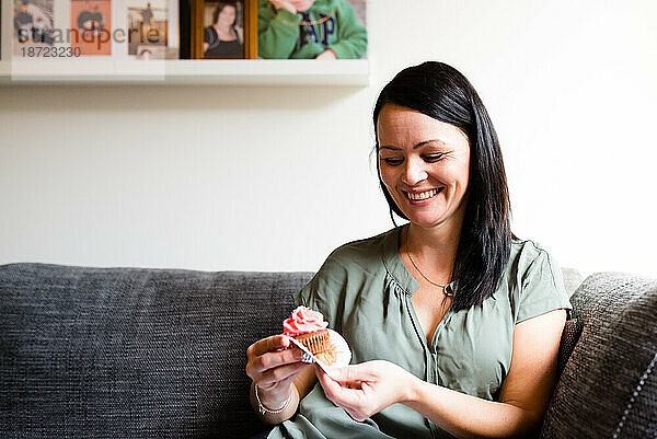 Eine Frau lächelt  während sie zu Hause auf dem Sofa einen Cupcake isst