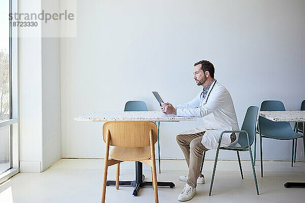 Seitenansicht eines männlichen Arztes  der einen Tablet-PC verwendet  während er auf einem Stuhl sitzt
