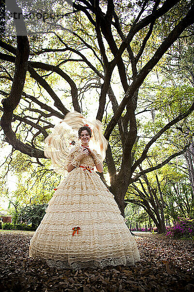 Eine junge Frau posiert in einem Southern Belle-Outfit  einschließlich eines großen  verzierten historischen Kleides und eines Sonnenschirms  unter einem Baum.