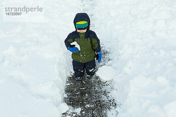 Kalter kleiner Junge geht mit einem Schneeball im Arm über einen geschaufelten Weg