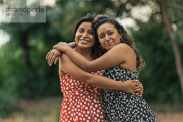 Ein Paar junge Latina-Frauen lächeln und umarmen sich auf einem Feld.