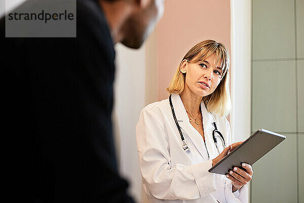 Blonde Ärztin hält Tablet-PC in der Hand  während sie männlichen Patienten betrachtet