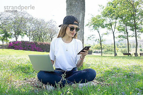 Vorderansicht einer glücklichen Studentin  die mit Laptop und Smartphone in einem grünen Park eines Universitätscampus arbeitet