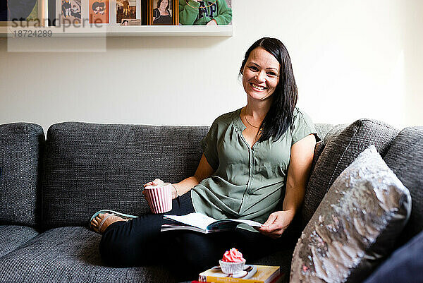 Eine Frau lächelt und liest auf dem Sofa mit einem Cupcake