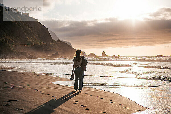 Junge Frau spaziert bei Sonnenuntergang an einem schwarzen Sandstrand