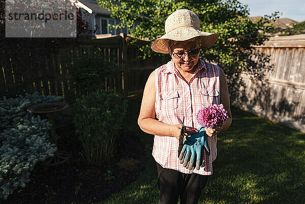 Ältere Frau mit Hut hält einen Strauß frisch geschnittener Blumen in einem Garten.