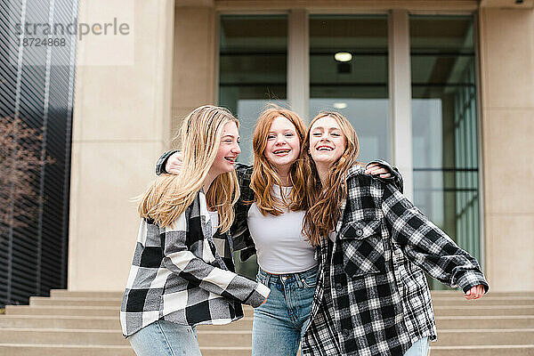 Drei lachende Teenager-Mädchen umarmen sich draußen auf Stufen.