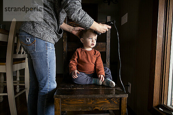 Mutter schneidet kleinen Jungen drinnen die Haare und sitzt auf einem Stuhl neben dem Fenster