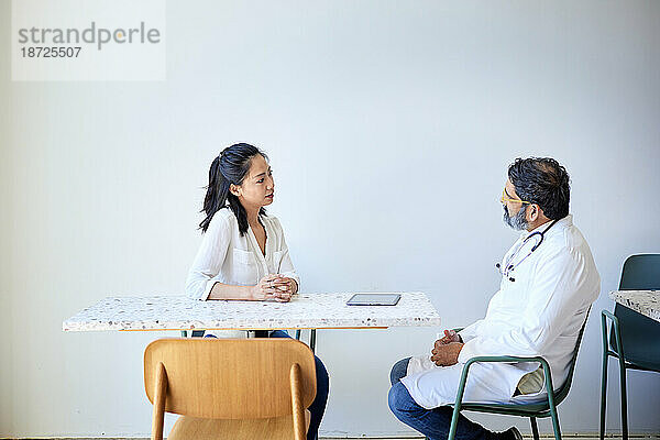 Patientin diskutiert mit männlichem Arzt  der auf einem Stuhl in der Klinik sitzt