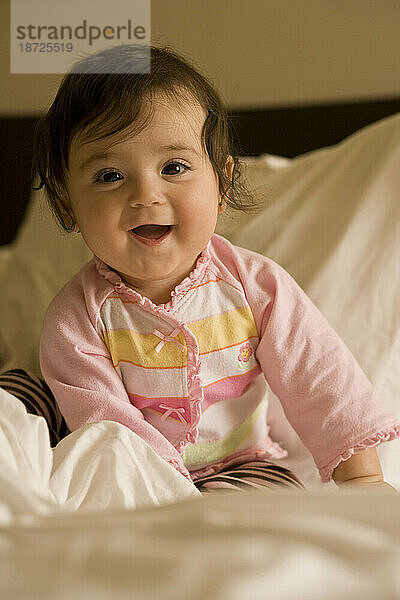 Ein glückliches kleines Mädchen sitzt auf einem Bett  umgeben von weichen weißen Laken und lächelt in die Kamera.